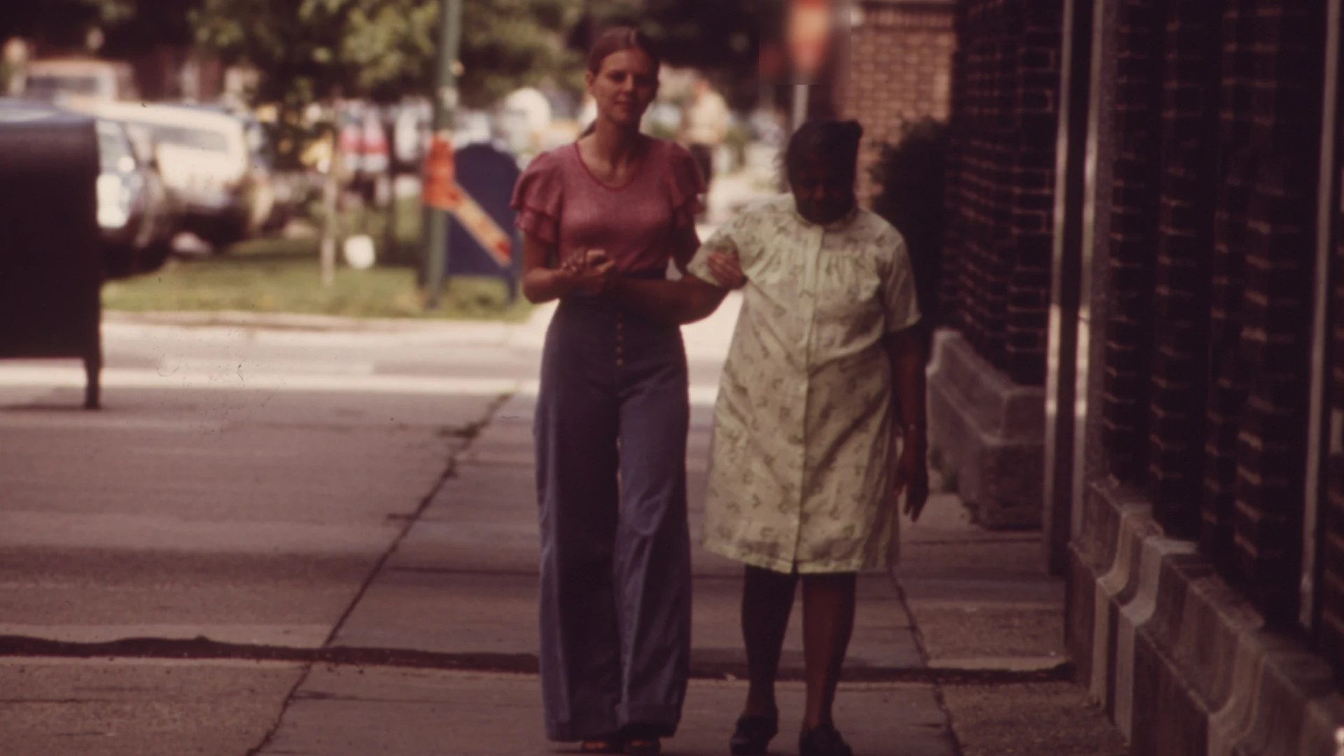 An older nursing home resident walking on a city sidewalk with help