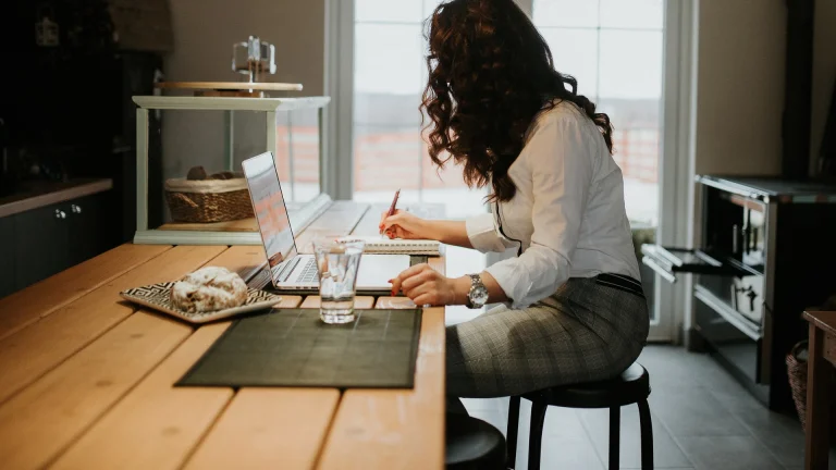 Young woman working on a laptop and writing notes at a kitchen table